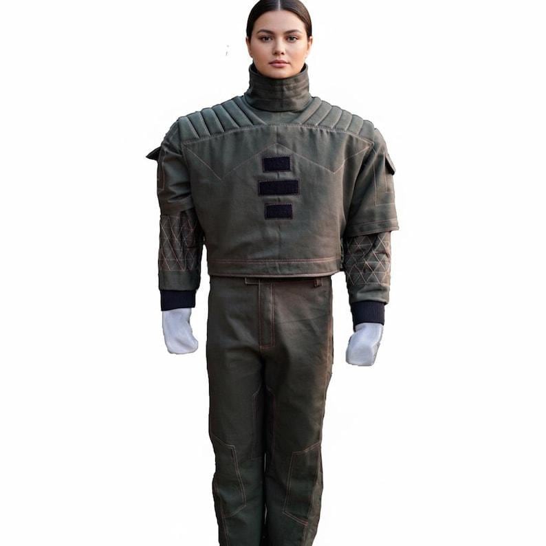May include: Olive green jumpsuit with a high collar and quilted shoulder detailing. The jacket has three black rectangular patches and the sleeves have a diamond pattern. The pants have red stitching. The model is wearing white gloves.