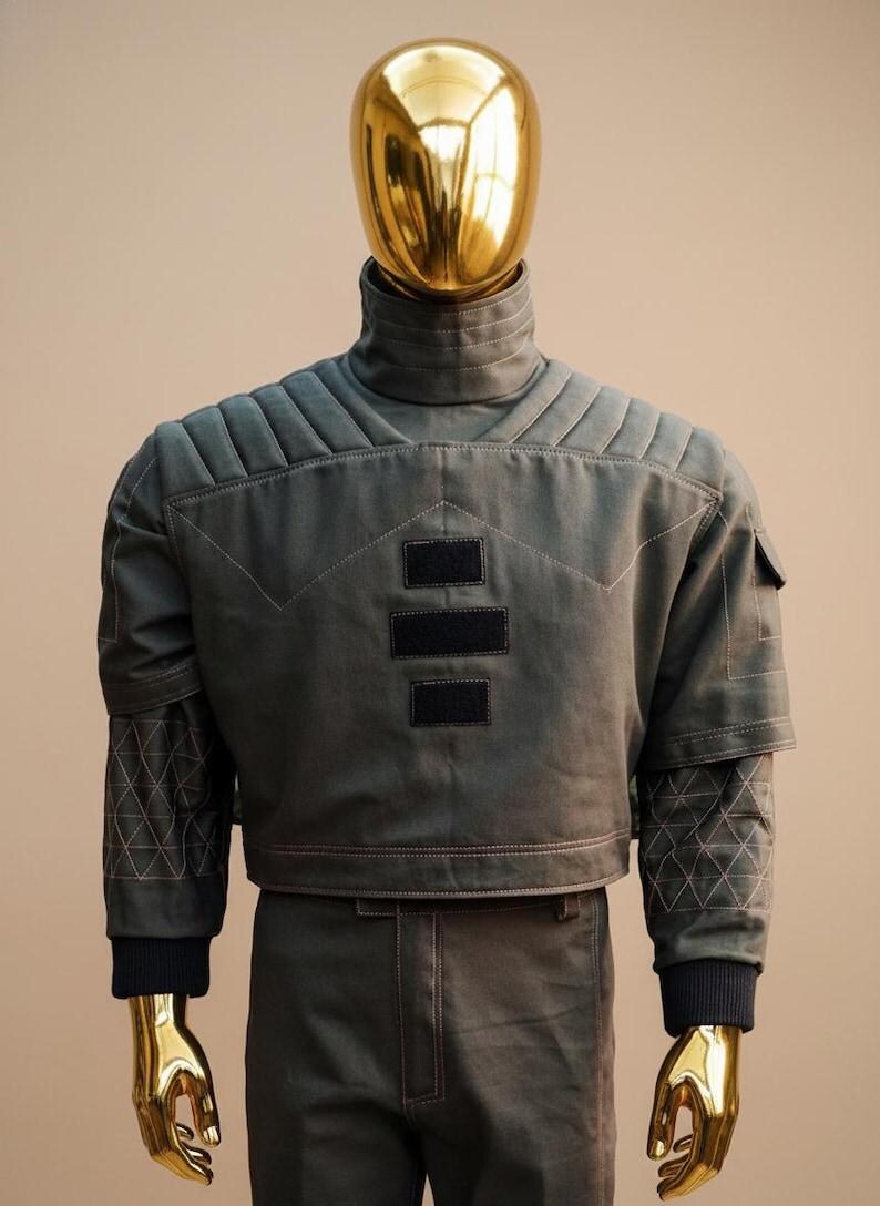 May include: A futuristic-style outfit on a mannequin with a gold head and hands. The ensemble includes a gray jacket with quilted shoulders, black patches, and a high collar, paired with matching pants. The jacket has geometric patterned sleeves.