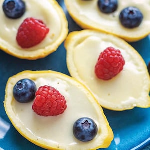 May include: Close-up of several lemon halves filled with a creamy custard and topped with fresh blueberries and raspberries. The lemons are yellow, the custard is pale yellow, and the berries are red and blue. The food is arranged on a blue plate.