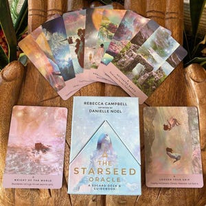 May include: The Starseed Oracle deck and guidebook by Rebecca Campbell and Danielle Noel. The deck includes 53 cards with colorful illustrations and text. The cards are fanned out on a wooden surface, with the guidebook in the center.
