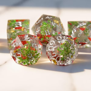 May include: Set of seven clear resin dice with embedded red and green botanical elements. The dice have silver numbering and are in various geometric shapes, including a d20, d6, and d4. The dice are on a white surface.