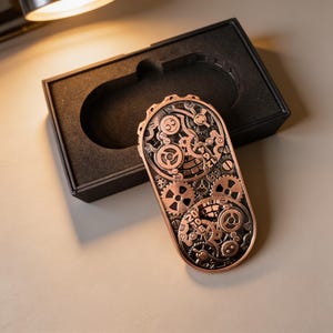 May include: A copper-colored, steampunk-style lighter with intricate gear designs. The lighter is rectangular with rounded ends and is displayed in a black box with a fitted interior. The gears are detailed and create a mechanical aesthetic.