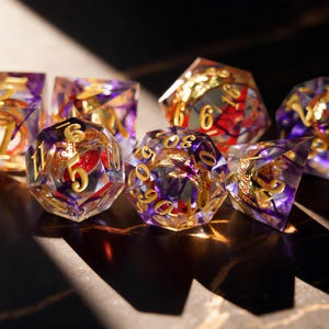 May include: A collection of polyhedral dice with a transparent resin body, featuring purple and gold accents. The dice have gold numerals and are designed for tabletop gaming. The set includes various shapes, including a D20, D12, and D6.