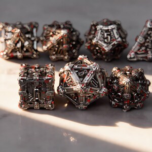 Könnte beinhalten: Ein Set aus sieben polyedrischen Würfeln mit einer komplexen, metallisch silbernen käfigartigen Struktur mit dunkelgrauen und roten Akzenten. Die Würfel haben komplizierte geometrische Designs und deutlich sichtbare Zahlen, angeordnet in zwei Reihen.
