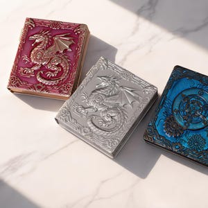 May include: Three rectangular cases with hinged lids. The left case is metallic pink with a raised dragon relief. The center case is metallic silver with a raised dragon relief. The right case is metallic blue with a circular gear and clockwork design.