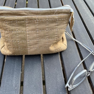 May include: A tan, rectangular cosmetic bag with a white zipper and stitching detail. Clear-framed eyeglasses rest on a wooden surface next to the bag. The bag has a quilted appearance.