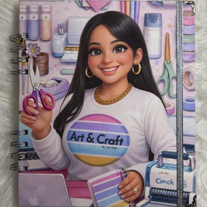 May include: A spiral-bound notebook with a colorful illustration of a woman surrounded by art supplies. The woman wears a white shirt with the text "Art & Craft by Astrid". Scissors and a notebook are visible.
