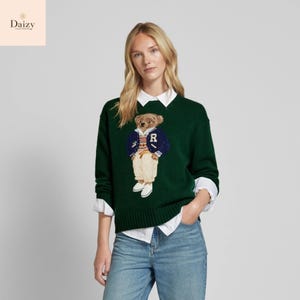 May include: A dark green sweater with a teddy bear motif, wearing a blue jacket and a white shirt. The bear's jacket has an "R" on it. The sweater is worn with blue jeans.