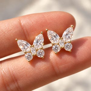 May include: A pair of gold-toned stud earrings shaped like butterflies. Each earring features clear, faceted, marquise-cut and round-cut stones arranged to form the wings and body of the butterfly, set in a polished gold-colored metal frame.
