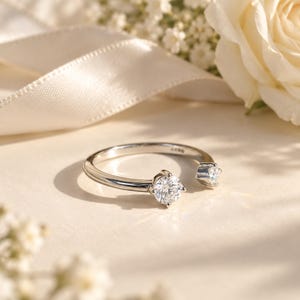May include: A silver-toned open ring featuring two round, clear, faceted stones set in four-prong settings at each end of the band. The band has a smooth, polished finish and includes a small hallmark stamp visible on the inner surface.