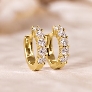 May include: A pair of gold hoop earrings featuring a row of five clear, round-cut stones set along the front curve of each hoop. The earrings have a polished metallic finish and a hinged closure mechanism visible on the side of the hoops.