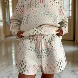 May include: A quilted two-piece set featuring a patchwork design. The set comprises a long-sleeved sweatshirt and shorts. The fabric is white with a pattern of Christmas trees, candy canes, and other festive elements. The shorts have a drawstring waist.