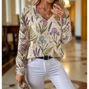 May include: Cream-colored V-neck sweater with a floral pattern featuring lavender, irises, and ferns in shades of purple, blue, and green. The sleeves have a unique open knit design. The sweater is paired with white pants and a black belt.