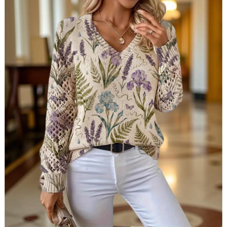 May include: Cream-colored V-neck sweater with a floral pattern featuring lavender, irises, and ferns in shades of purple, blue, and green. The sleeves have a unique open knit design. The sweater is paired with white pants and a black belt.
