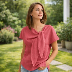 May include: A coral-colored short-sleeved top with a unique bow detail at the neckline. The top has a relaxed fit and is made of a lightweight fabric. The model is wearing blue jeans and is standing outside in a garden setting.