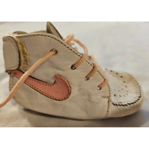 May include: A high-top baby shoe crafted from off-white leather, featuring a pink leather swoosh logo on the side, peach-colored laces, and a perforated toe box. The ankle collar includes a textured brown patch, and the sole is stitched.