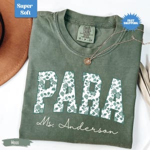 May include: A moss green t-shirt with the word "PARA" in large, floral-patterned letters. Below, the name "Ms. Anderson" is written in cursive. The shirt has a crew neck and a small Comfort Colors tag. A gold necklace with a pendant is draped on the shirt.