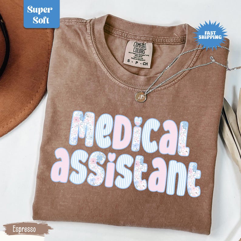 May include: Espresso-colored t-shirt with the words "MEDICAL ASSISTANT" in a floral and striped design. The shirt has a crew neck and short sleeves. A silver necklace with a pendant is draped across the shirt.