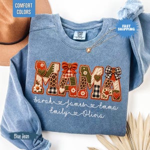 May include: A blue denim-coloured crewneck sweatshirt featuring the word MAMA in large, patchwork-style letters with various patterns including leopard print, plaid, and floral. Below the text, the names Sarah, James, Emma, Emily, and Olivia are embroidered in white script.
