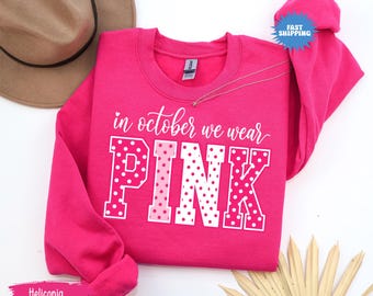 May include: A bright pink long-sleeve crewneck sweatshirt featuring the white text "in october we wear" above the word "PINK" in large block letters. Each letter of the word "PINK" is decorated with a white polka dot pattern on a pink background.