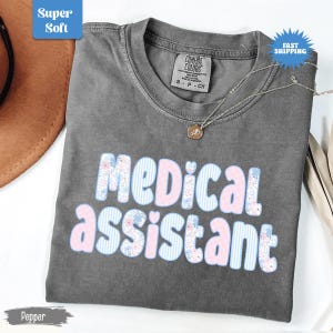 May include: A gray t-shirt with the words "MEDICAL ASSISTANT" in a pastel floral and striped design. The shirt has a crew neck and short sleeves. A silver necklace with a small pendant rests on the shirt.