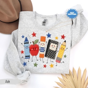 May include: A light gray crewneck sweatshirt featuring a colorful embroidered design of anthropomorphic school supplies including a blue crayon, a red apple, an orange glue stick, a black notebook, and a yellow pencil surrounded by small stars.