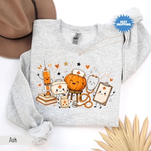 May include: A light gray crewneck sweatshirt featuring a graphic print of a smiling pumpkin wearing a nurse cap, a syringe, a medical kit, a pill bottle, a stethoscope, and a clipboard, all with cartoon faces, surrounded by orange hearts and stars.