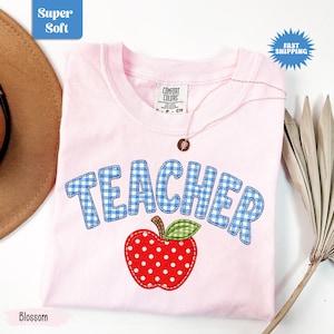 May include: A light pink t-shirt featuring the word TEACHER in blue and white gingham letters arched above a red apple graphic with white polka dots and a green gingham leaf. The shirt has a crew neckline and a Comfort Colors brand label.
