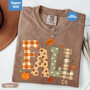 May include: An espresso-coloured short-sleeve t-shirt featuring the word FALL in large block letters. Each letter has a different pattern: orange plaid, floral print, sage green polka dots, and brown plaid. Small pumpkin and leaf graphics surround the text.