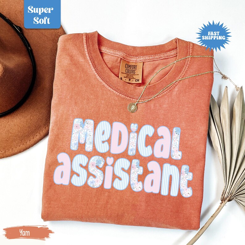 May include: A yam-colored t-shirt with the words "MEDICAL ASSISTANT" in a playful font. The letters are decorated with pink, blue, and floral patterns. A gold necklace and a Comfort Colors tag are visible. The shirt is labeled "Super Soft" and "Fast Shipping".