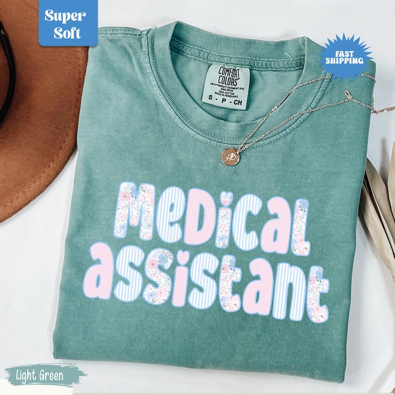 May include: A light green t-shirt with the words "Medical Assistant" in a floral and striped design. The shirt has a crew neck and short sleeves. A silver necklace with a pendant is draped across the shirt. The shirt is labeled "Super Soft" and "Fast Shipping".
