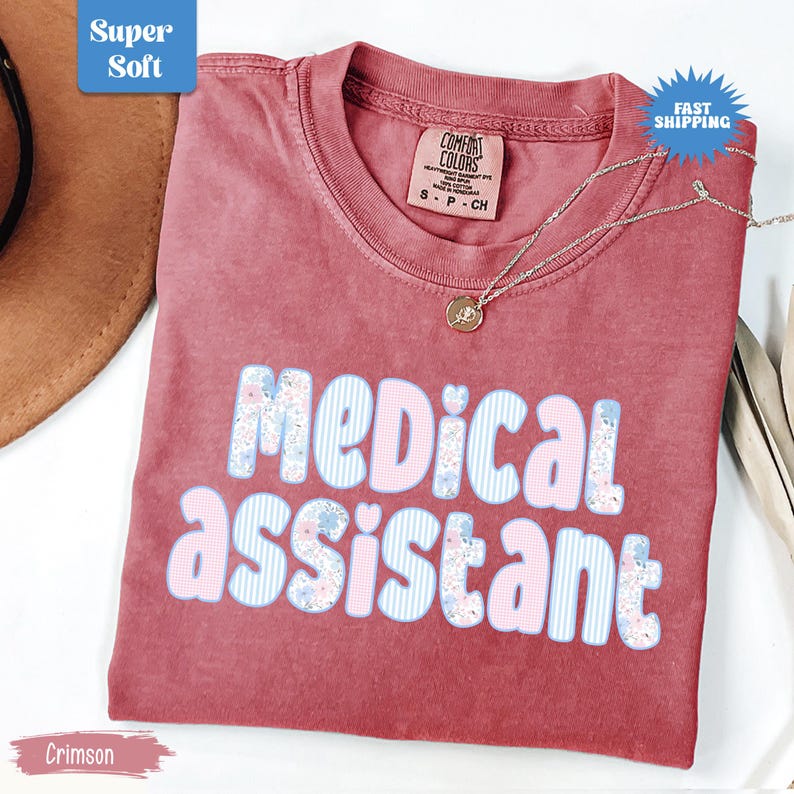 May include: Crimson-colored t-shirt with the words "MEDICAL ASSISTANT" in a playful font. The letters are decorated with blue stripes and floral patterns. A silver necklace with a pendant rests on the shirt. The shirt has a tag that says "Comfort Colors".
