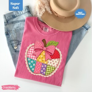 May include: A pink t-shirt featuring a large apple-shaped applique made of various patterned fabric patches including gingham, floral, and polka dot prints. The apple is accented with a pink bow and a green leaf, all bordered by white lace.