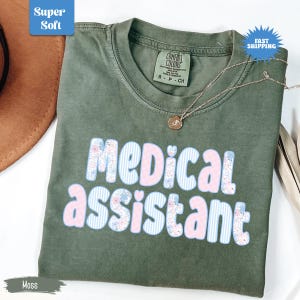 May include: Moss green t-shirt with the words "MEDICAL ASSISTANT" in a floral and striped design. The shirt has a crew neck and short sleeves. A silver necklace with a pendant is draped on the shirt.