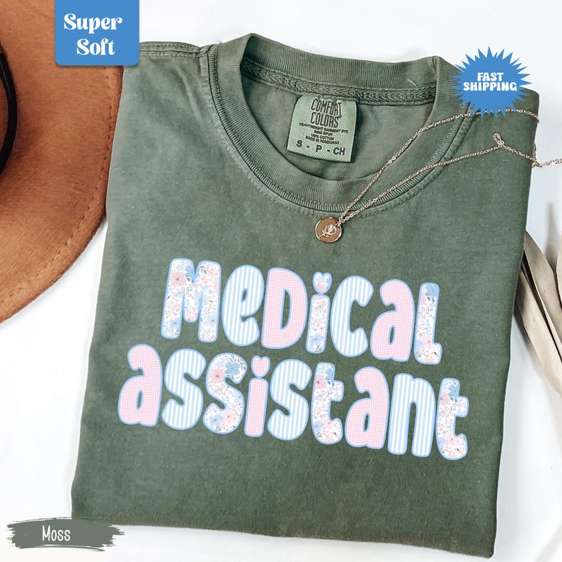 May include: Moss green t-shirt with the words "MEDICAL ASSISTANT" in a floral and striped design. The shirt has a crew neck and short sleeves. A silver necklace with a pendant is draped on the shirt.