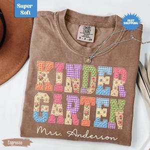 May include: Espresso-coloured t-shirt with the word "KINDERGARTEN" in colourful patchwork letters. The name "Mrs. Anderson" is below the word. A silver necklace with a pendant is on the shirt. The shirt has the words "Super Soft" and "Fast Shipping" on it.