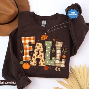 May include: A dark chocolate brown long-sleeve sweatshirt featuring the word FALL in large, patterned block letters. The letters include orange gingham, floral, sage green polka dot, and plaid prints, accented with small pumpkin and leaf graphics.