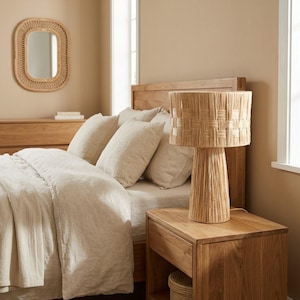 Puede incluir: Una lámpara de mesa con base cilíndrica y pantalla de tambor, ambas fabricadas con fibras naturales trenzadas en color beige claro. La lámpara se apoya sobre una mesita de noche de madera junto a una cama con ropa de cama beige y cabecero.