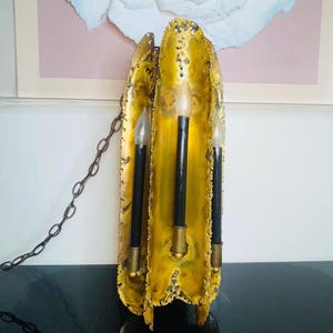 May include: A vintage gold-toned wall sconce with three faux candle lights. The sconce has a textured, organic shape and a dark chain for hanging. The candle lights have black bases and clear, flame-shaped bulbs. The sconce is mounted on a wall.