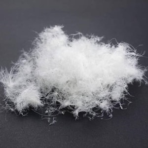 Op de afbeelding: Een close-up van een hoopje zachte, witte donsveren. De veren zijn zacht en delicaat, met een ietwat ijle textuur. De dons ligt op een donkergrijze achtergrond, wat de helderwitte kleur van de veren benadrukt.