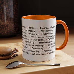May include: White ceramic mug with an orange interior and handle. The mug is printed with a list of words. A silver spoon and a macaron are on a wooden surface next to the mug.
