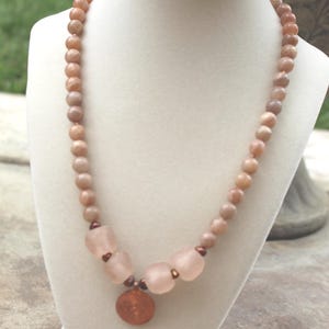 Puede incluir: Un collar con una hebra de pequeñas cuentas redondas de color melocotón. El collar tiene cuentas rosas esmeriladas más grandes y un colgante de color cobre. El collar se exhibe en un busto de joyería blanco.