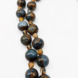 May include: A necklace featuring two strands of round beads with a marbled blue and metallic bronze finish, interspersed with smaller, translucent amber-colored faceted beads, creating a textured and reflective appearance throughout the piece.