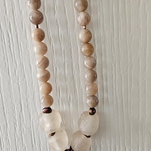 May include: A necklace featuring a strand of light brown, spherical beads. The necklace has a pendant with a copper spiral design and several larger, irregular-shaped, clear beads. The beads are strung on a delicate cord.