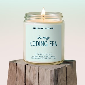 May include: A lit glass jar candle with a light blue label that reads "Fireside Stories" and "in my CODING ERA". The candle is labeled "Lychee Lotus" and is hand-poured in the USA. The candle is 9 oz (255 g).