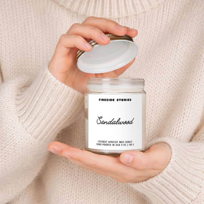 Pode incluir: Um frasco de vela de vidro transparente com um r&oacute;tulo branco contendo o texto FIRESIDE STORIES, Sandalwood e COCONUT APRICOT WAX CANDLE HAND-POURED IN USA 9 OZ | 198 G. A tampa de metal prateado est&aacute; aberta, revelando a cera branca.