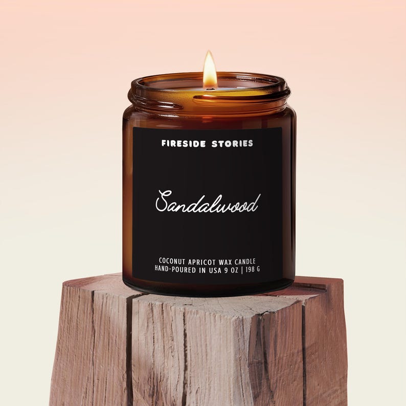 May include: A lit, brown glass jar candle with a black label that reads "FIRESIDE STORIES" and "Sandalwood." The label also states it is a coconut apricot wax candle, hand-poured in the USA, 9 oz (255 g). The candle sits on a wooden block.
