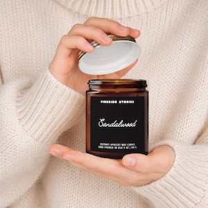 May include: An amber glass candle jar with a black label featuring the text Fireside Stories and Sandalwood. The label specifies Coconut Apricot Wax Candle Hand-Poured in USA 9 oz | 198 g. The jar has a silver metal lid being held open.