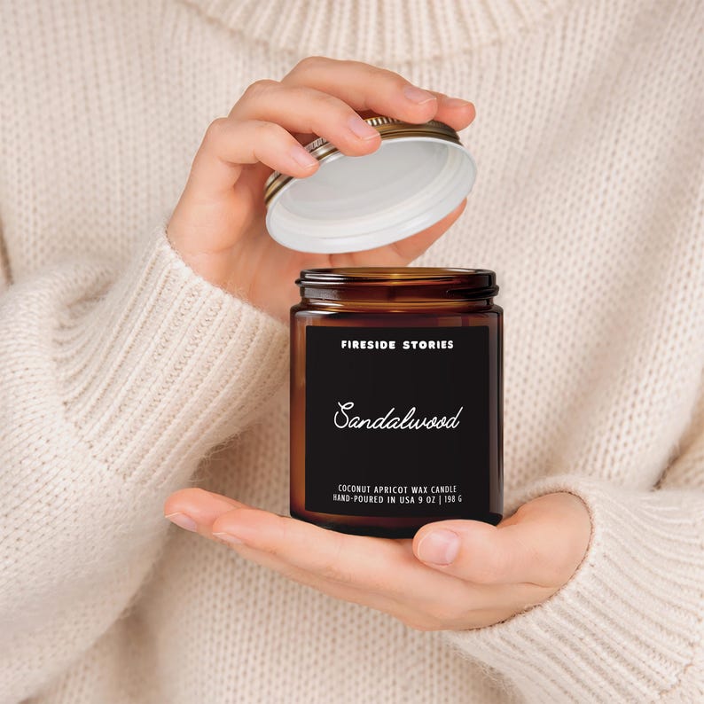 May include: An amber glass candle jar with a black label featuring the text Fireside Stories and Sandalwood. The label specifies Coconut Apricot Wax Candle Hand-Poured in USA 9 oz | 198 g. The jar has a silver metal lid being held open.