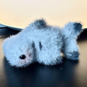 Puede incluir: Un pequeño juguete de peluche con forma de pez, de color gris claro y textura peluda. El pez presenta un solo ojo oscuro y redondo, una pequeña aleta oscura en la parte superior y una cola con detalles en gris más oscuro en las puntas.